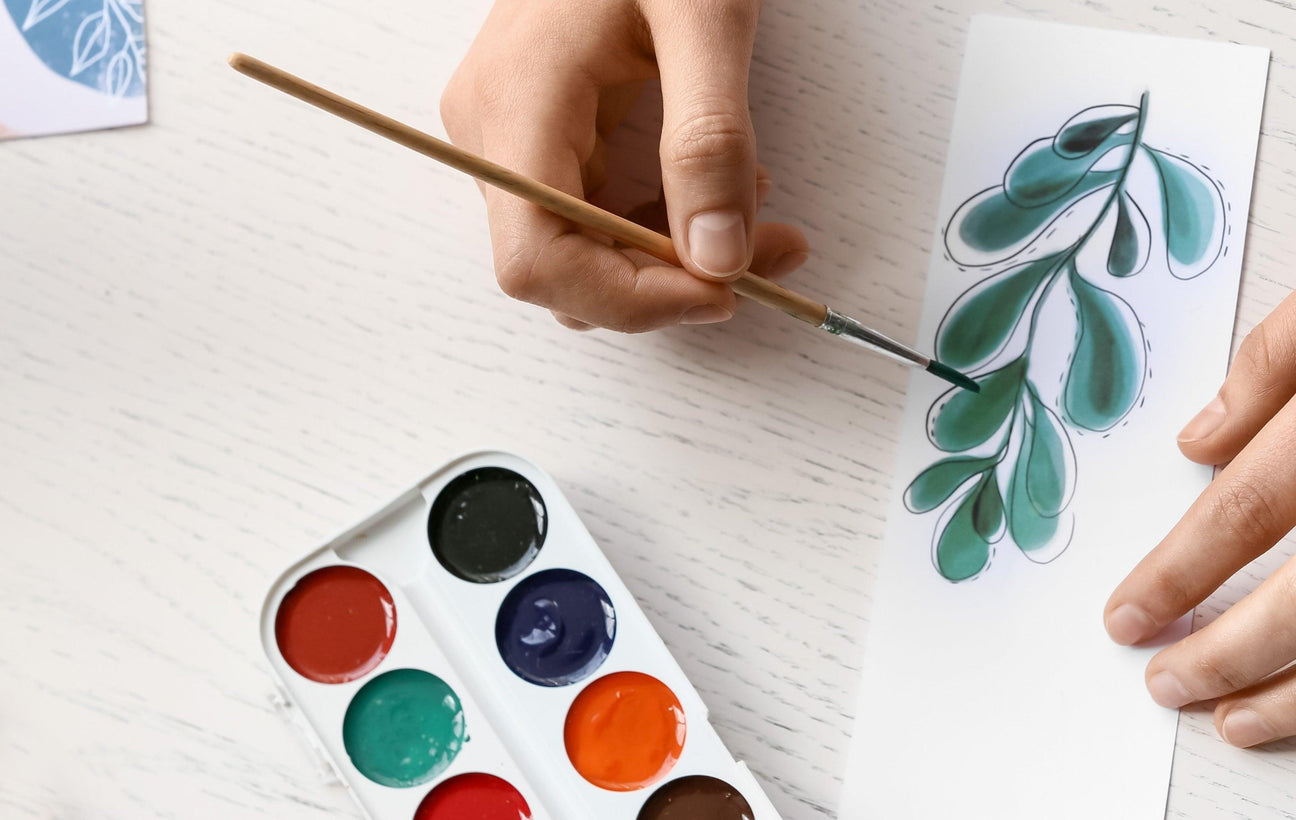 Hand painting a green leaf design with watercolors, showcasing diverse art supplies for creative projects.