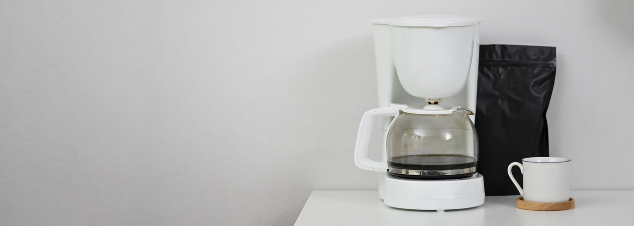 White coffee maker with glass carafe, black coffee bag, and white mug on a minimalist counter.