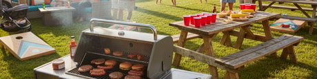 Outdoor summer cookout with a grill full of burgers and hot dogs, picnic tables, cornhole, and guests enjoying the sunny lawn.
