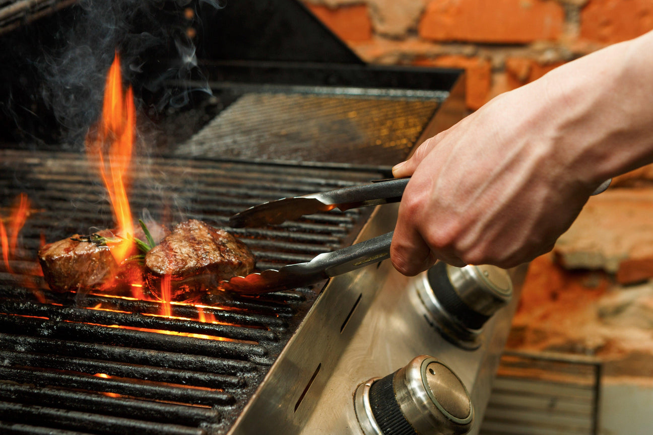 Hand using tongs to flip sizzling steaks on a gas grill with flames, ideal for B2B grilling supplies.