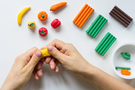 Hands molding yellow clay, surrounded by colorful clay fruits, vegetables, and bars for creative projects.
